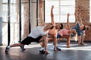 Eine Gruppe von fünf Personen führt gemeinsam Fitnessübungen in einem Fitnessstudio mit industriellem Loft-Charakter durch. Die Teilnehmer tragen bunte Sportkleidung und befinden sich alle in der gleichen Fitness-Position, wobei sie sich seitlich abstützen und einen Arm nach oben strecken.  Eine Gruppe von fünf Personen führt gemeinsam Fitnessübungen in einem Fitnessstudio mit industriellem Loft-Charakter durch. Die Teilnehmer tragen bunte Sportkleidung und befinden sich alle in der gleichen Fitness-Position, wobei sie sich seitlich abstützen und einen Arm nach oben strecken.
